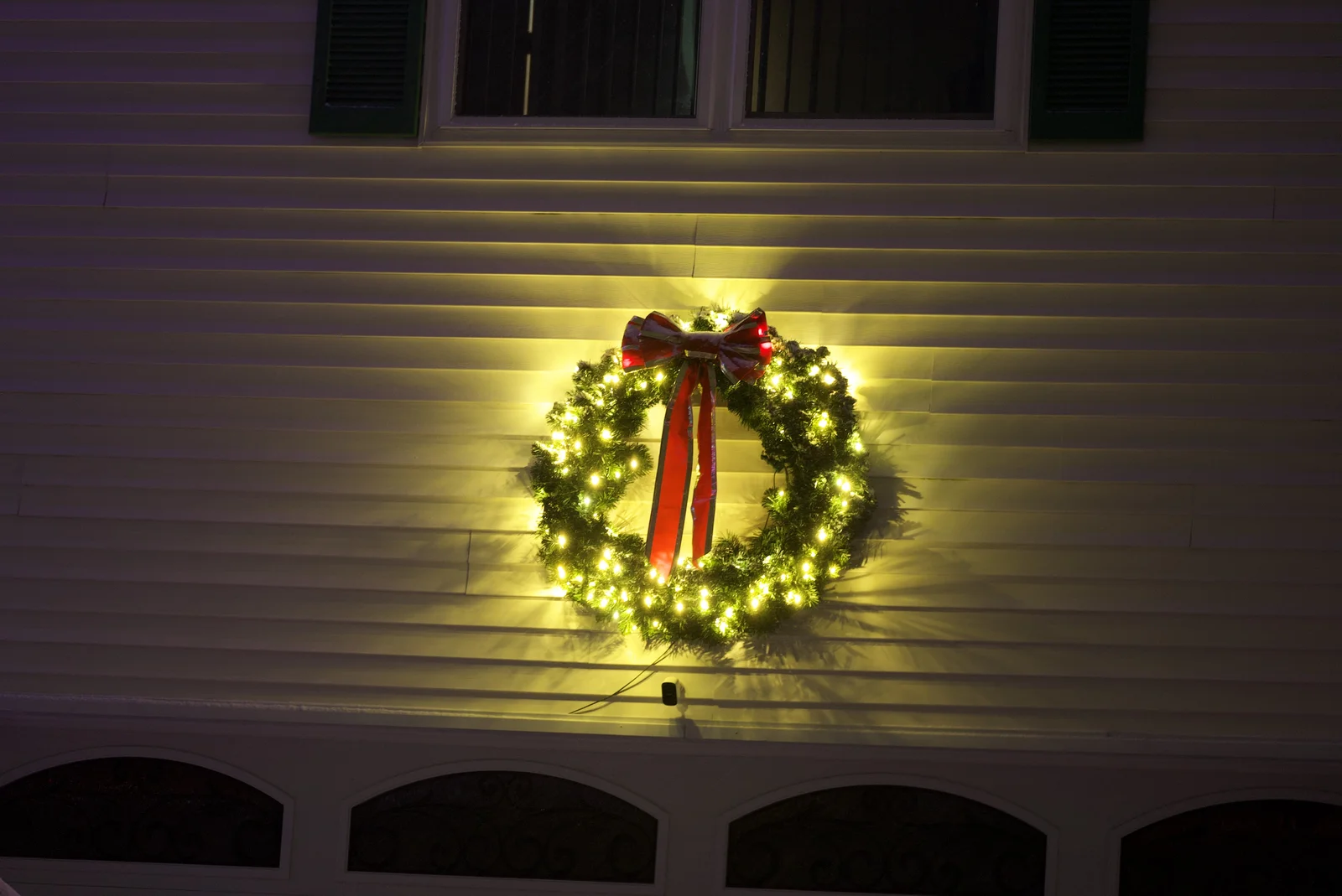 Green Acres holiday lighting installation featuring illuminated Christmas wreath with red bow on white house exterior