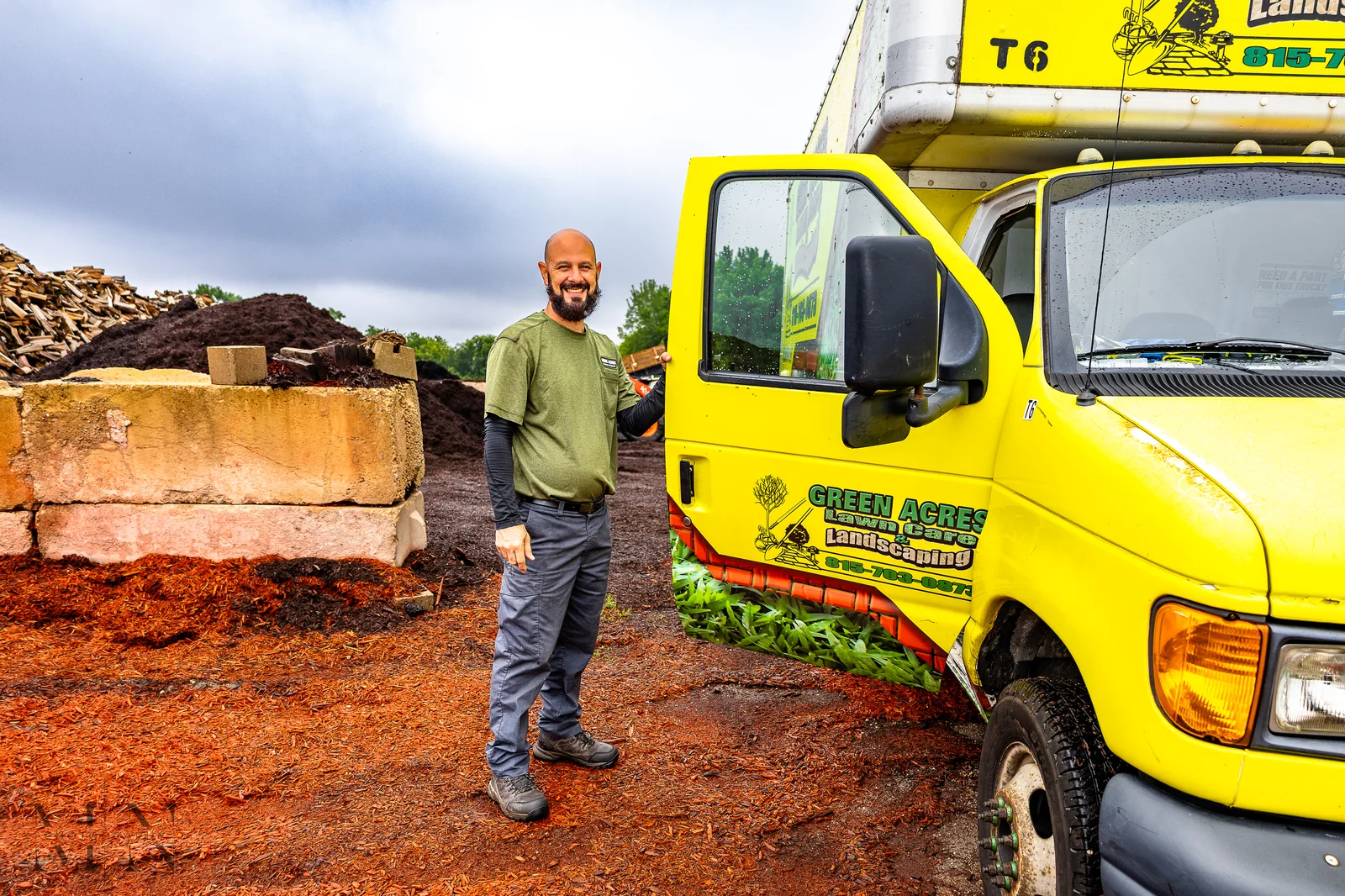 Green Acres Landscaping crew member standing beside branded yellow truck at mulch yard with bulk mulch products